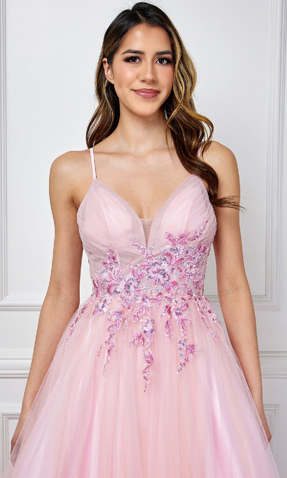 A woman stands smiling against a white paneled wall, wearing the Long Pastel Prom Ball Gown: Amelia Couture SU088—a light pink, sleeveless A-line gown with floral embellishments.